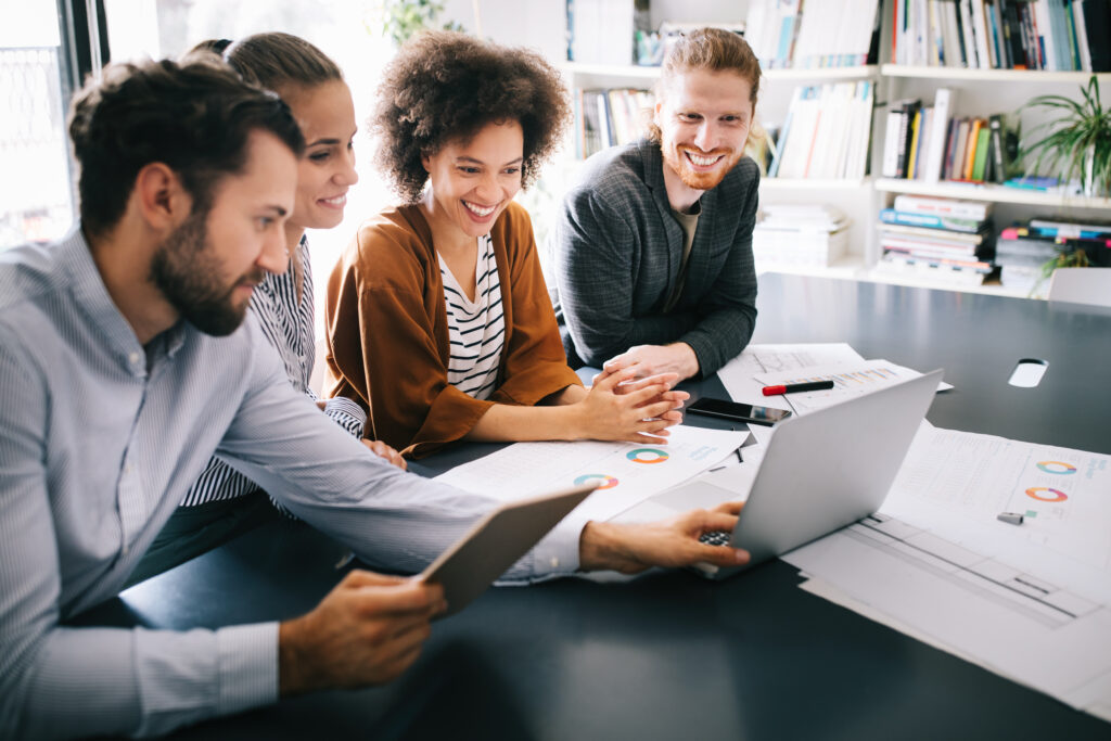 A diverse team of four people, male and female, smiling and sitting in front of an open laptop. Paperwork litters the desk.