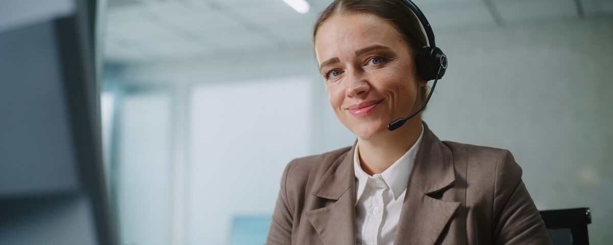 A smiling woman with blue eyes sitting in front of her computer, wearing a headset and a brown suit jacket.