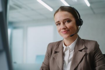 A smiling woman with blue eyes sitting in front of her computer, wearing a headset and a brown suit jacket.