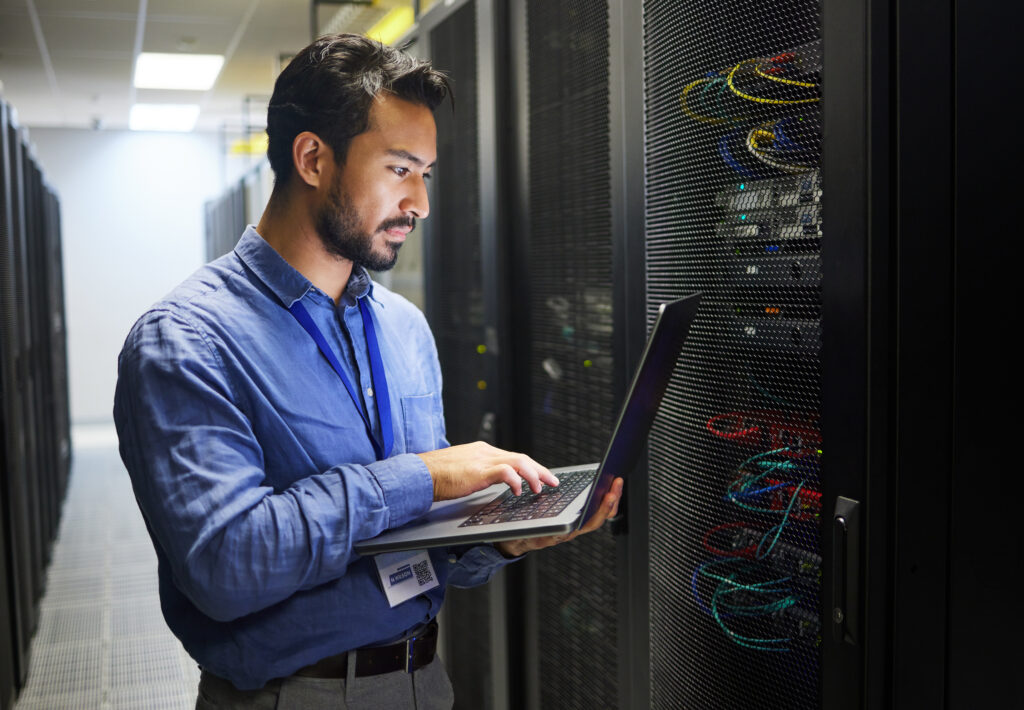 A man with a dark beard holds a laptop and stands in a server room. He is wearing a blue lanyard and a dress shirt.