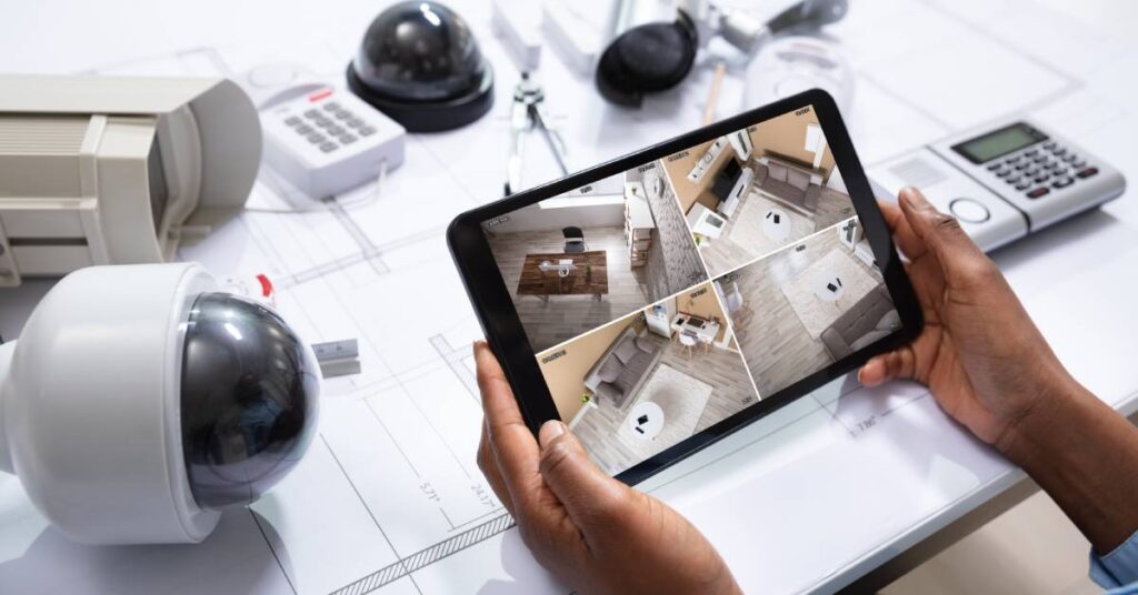 A person holding a tablet that displays four angles of a home interior. Multiple security cameras sit on the desk.