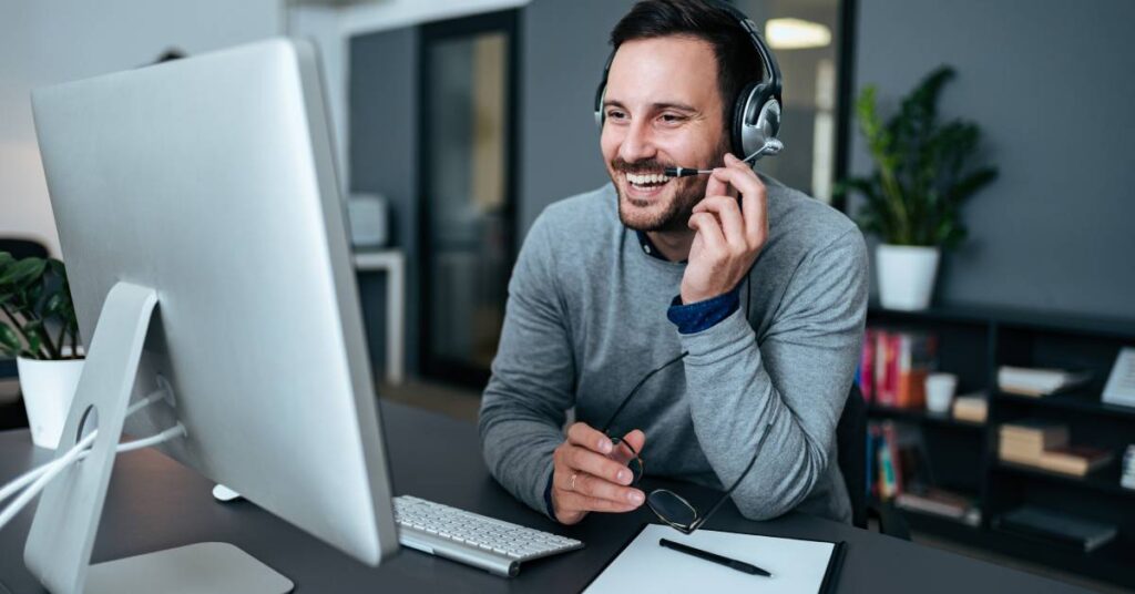 A man wearing a gray long-sleeved shirt sits at a desk with a monitor. He speaks on a headset microphone and smiles.