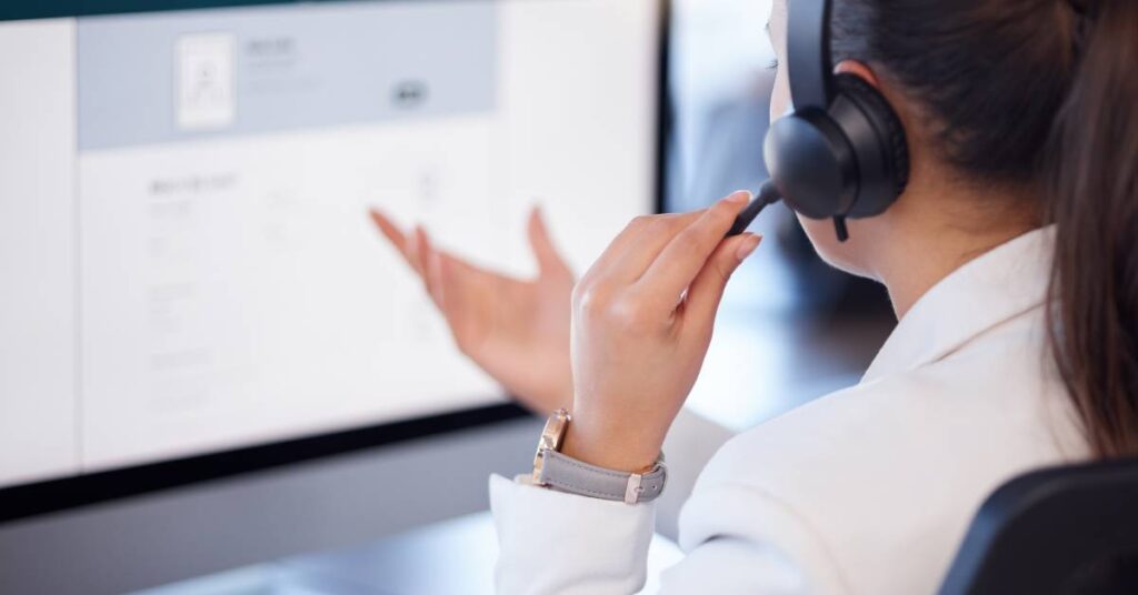 A woman with a ponytail and a headset gesturing at a computer in front of her. The perspective is over her shoulder.